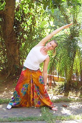 Dreieck Die Kursleiterin steht vor Palmen und macht eine stehende Yoga-Stellung, indem sie den rechten Arm nach schräg oben streckt und sich nach links beugt. Die Beine sind gespreizt und der linke Arm hängt nach unten. Diese Stellung nennt man Dreieck.