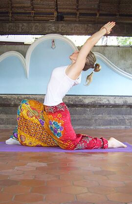 Yoga-Halle Das Bild zeigt die Yoga-Lehrerin in einer Yoga-Halle in Indien. Sie macht eine Yoga-Stellung, indem sie links auf dem Boden kniet und das rechte Bein nach vorne im 90-Grad-Winkel abstellt. Die Arme sind weit nach oben und hinten gestreckt.