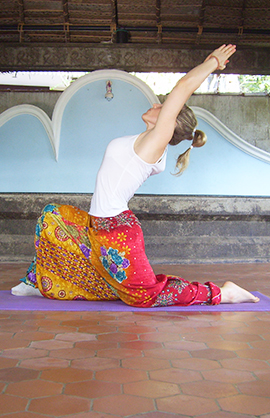 Das Bild zeigt die Yoga-Lehrerin in einer Yoga-Halle in Indien. Sie macht eine Yoga-Stellung, indem sie links auf dem Boden kniet und das rechte Bein nach vorne im 90-Grad-Winkel abstellt. Die Arme sind weit nach oben und hinten gestreckt.