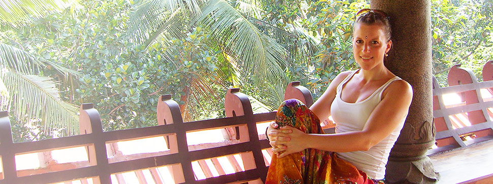 Die Yoga-Lehrerin sitzt ganz entspannt auf einer Terrasse und lächelt in die Kamera. Im Hintergrund sind Palmen.