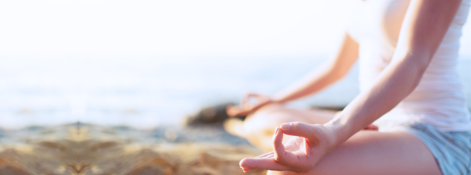 Das Bild zeigt eine Frau, die am Meer in einer Meditationshaltung sitzt. Die Beine sind gekreuzt, die Hände liegen auf den Knien, Zeigefinger und Daumen berühren sich.